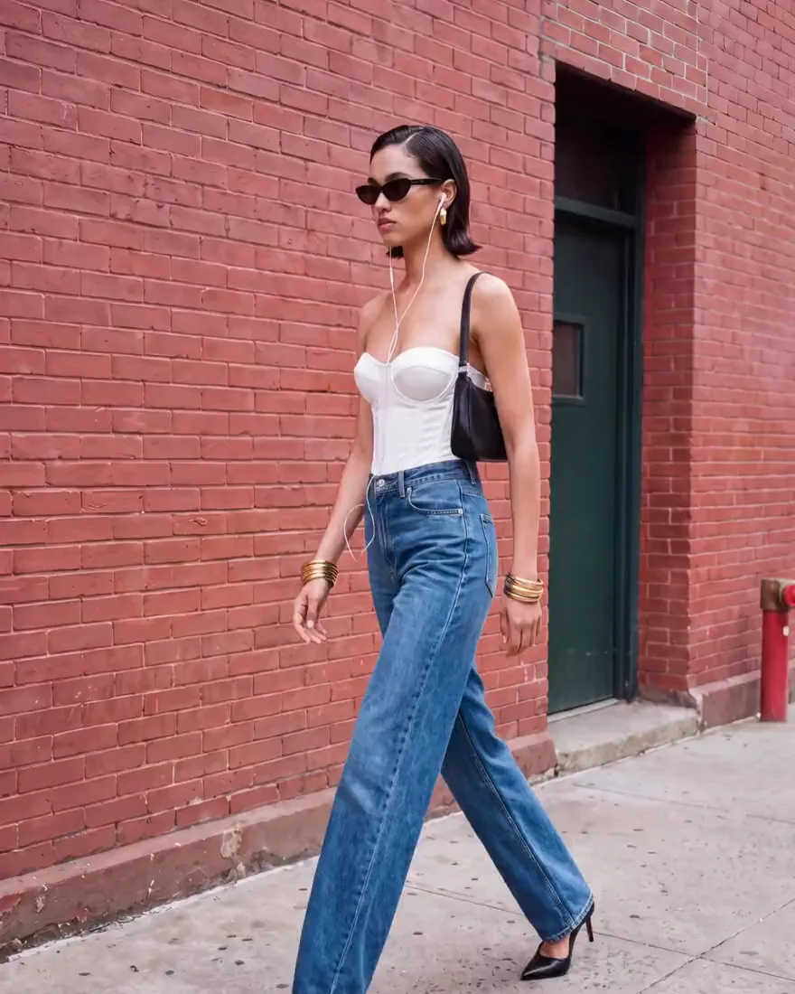 Ein KI-Model in weißem Korsett-Top und blauer Jeans läuft an einer Backsteinmauer vorbei, als Foto und als daraus erstelltes Kurzvideo